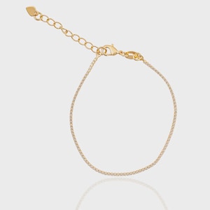 Peut inclure: Un bracelet doré délicat avec une rangée de petites pierres scintillantes. Le bracelet est doté d'une chaîne réglable et d'un charm en forme de cœur. Le fermoir est de type mousqueton. Le bijou est présenté sur une surface réfléchissante.