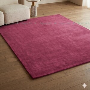 May include: A large, rectangular magenta rug dominates the foreground, set on a hardwood floor. The rug's texture appears soft and plush. A white armchair and a small side table with a marble base are visible in the background, suggesting a living room setting.