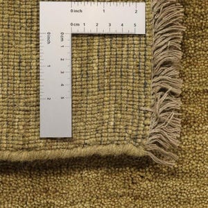 Op de afbeelding: Close-up van een beige tapijt met een franje rand. Een liniaal is op het tapijt gelegd om de textuur en de weving te laten zien. De liniaal meet in inches en centimeters.