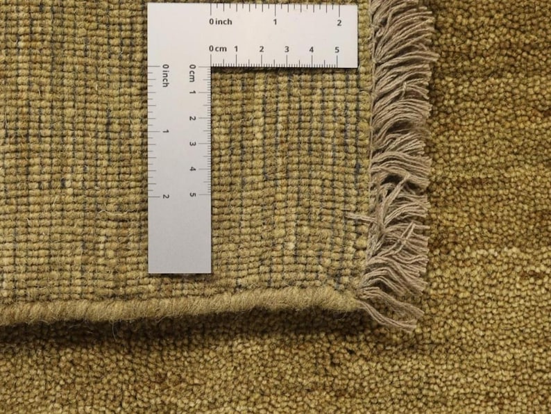 Op de afbeelding: Close-up van een beige tapijt met een franje rand. Een liniaal is op het tapijt gelegd om de textuur en de weving te laten zien. De liniaal meet in inches en centimeters.