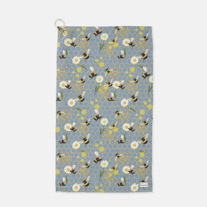 May include: A blue golf towel with a repeating pattern of bees, flowers, and honeycomb. The towel has a white border and a small logo in the bottom right corner.