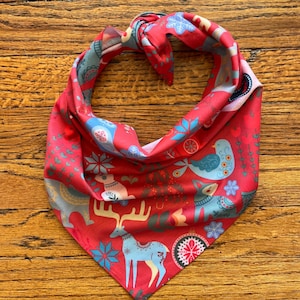 May include: Red bandana with a Scandinavian folk art pattern featuring animals, birds, and geometric designs. The bandana is folded into a triangle shape.
