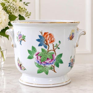 May include: A white ceramic planter with two side handles and a gold-trimmed rim. The exterior features hand-painted floral motifs in orange, pink, and green, along with colorful butterfly illustrations scattered across the surface.