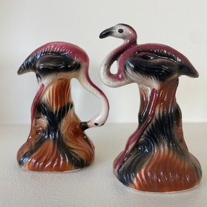 May include: A pair of ceramic flamingo salt and pepper shakers. The shakers are painted in shades of pink, brown, and black. The flamingo on the left is bending its head down, while the flamingo on the right is standing upright.