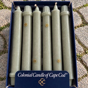 May include: A set of six sage green taper candles arranged in a dark blue box. Each candle features a ribbed top and a small gold circular emblem with the letters CC. The box is labeled Colonial Candle of Cape Cod with a gold CC logo at the bottom.