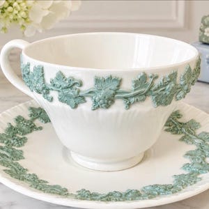 May include: A white ceramic teacup and saucer set featuring a raised, light green relief pattern of grapevines and leaves encircling the exterior of the cup and the rim of the saucer. The cup has a curved handle and a smooth, glossy finish.