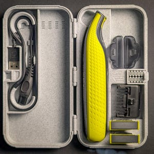 May include: An open, light gray travel case containing a yellow and gray electric shaver, charging cable, and various trimmer attachments. The shaver has a textured grip. The case is on a dark gray surface.