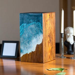 Pode incluir: Caixa decorativa retangular de madeira com um design de onda de resina azul e branca em um lado. A madeira tem um tom marrom quente. A caixa está em uma mesa de madeira com velas e uma pequena moldura.