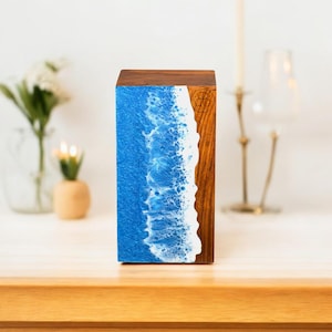 Könnte beinhalten: Ein rechteckiger Holzblock mit einem blau-weißen Harzdesign, das Meereswellen nachahmt. Die rechte Seite und die Oberseite sind aus Naturholz. Ein kleines Logo ist in das Holz geschnitzt. Der Block steht aufrecht.