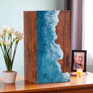 Op de afbeelding: Een houten urn met een blauw en wit oceaan golf ontwerp aan de voorkant. De rechthoekige urn staat op een houten oppervlak. Een kleine ingelijste foto en een kaars zijn in de buurt.