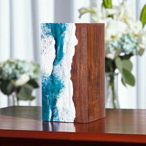 Könnte beinhalten: Ein rechteckiger Holzblock mit einem einzigartigen Design. Eine Seite zeigt ozeanische Wellen in Blau und Weiß, während die andere aus dunklem, braunem Holz besteht. Der Block wird auf einer Holzoberfläche präsentiert, was einen Kontrast erzeugt.