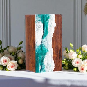 Könnte beinhalten: Eine Holzbox mit einem türkis-weißen Harzdesign, das Meereswellen nachahmt. Die Box hat eine dunkelbraune Holzmaserung. Das Harzdesign ist ein vertikaler Streifen. Weiße Rosen sind im Vordergrund arrangiert.
