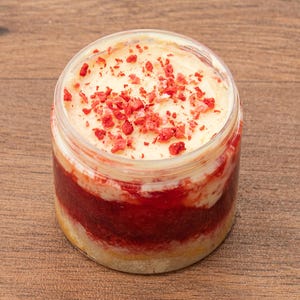 May include: A layered dessert in a clear jar. The dessert has a light-coloured top with red sprinkles, a red middle layer, and a light-coloured bottom layer. The jar is on a wooden surface.