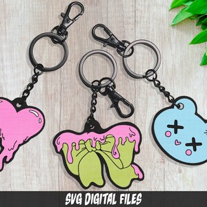 May include: Three keychains with different designs: a pink melting heart, a green bow with pink dripping icing, and a light blue bear with pink hearts and black crosses.  SVG DIGITAL FILES