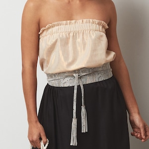 May include: A white snakeskin belt with tassels, worn over a cream-colored strapless top and a black skirt.