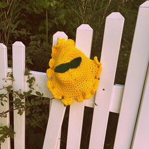 Pu&ograve; includere: Un cappello di uncinetto giallo con occhi di feltro verde e baffi di feltro nero. Il cappello &egrave; appoggiato su una staccionata bianca.
