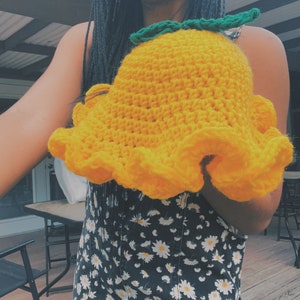 Pu&ograve; includere: Un cappello a secchiello di uncinetto giallo con foglie verdi in cima. Il cappello &egrave; tenuto da una persona che indossa un vestito floreale bianco e nero.