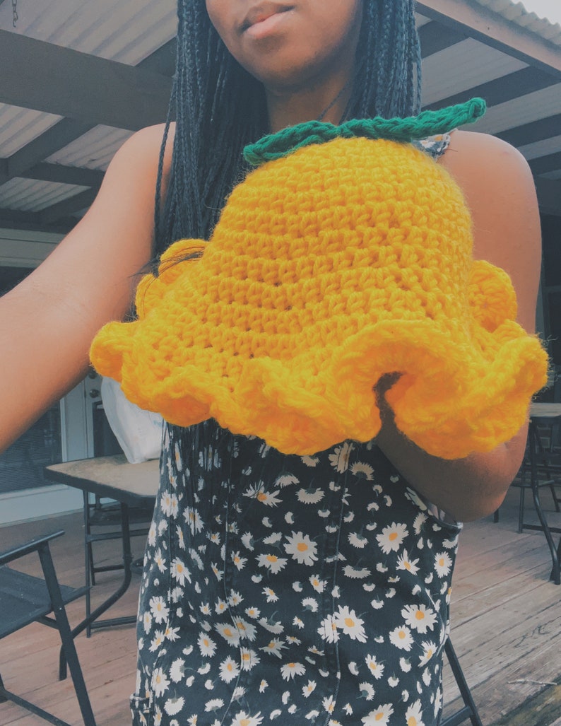 Pu&ograve; includere: Un cappello a secchiello di uncinetto giallo con foglie verdi in cima. Il cappello &egrave; tenuto da una persona che indossa un vestito floreale bianco e nero.