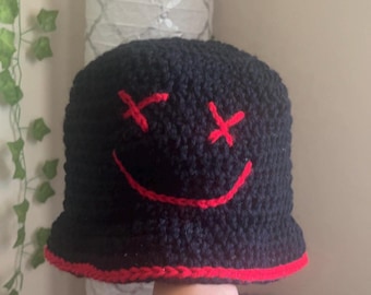 Può includere: Cappello a secchiello nero con un sorriso ricamato rosso con X per gli occhi.