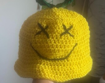 Può includere: Un cappello all'uncinetto giallo brillante fatto a mano con un design a faccina sorridente. Il cappello presenta occhi cuciti neri e una bocca curva, con foglie verdi sulla parte superiore. Un accessorio unico e divertente.