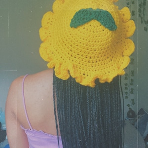 Pu&ograve; includere: Un cappello di girasole in crochet giallo con un baffo in crochet verde. Il cappello ha un bordo arricciato ed &egrave; indossato da una persona con lunghe trecce nere.