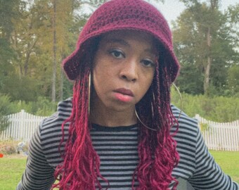 Può includere: Una persona con i capelli rosa indossa un cappello a secchiello in crochet bordeaux, una camicia a maniche lunghe a righe bianche e nere e jeans blu strappati. La persona è seduta su una sedia pieghevole nera davanti a una recinzione bianca.
