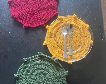 Può includere: Tre tovagliette all'uncinetto in rosso, giallo e verde. Ogni tovaglietta ha un design circolare con un bordo rialzato e un centro testurizzato. Le tovagliette sono su una superficie nera.