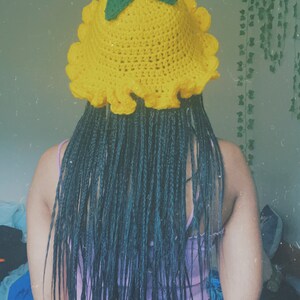Pu&ograve; includere: Un cappello a secchiello di uncinetto giallo con foglie verdi in cima. Il cappello ha un bordo arricciato ed &egrave; indossato da una persona con lunghe trecce nere.