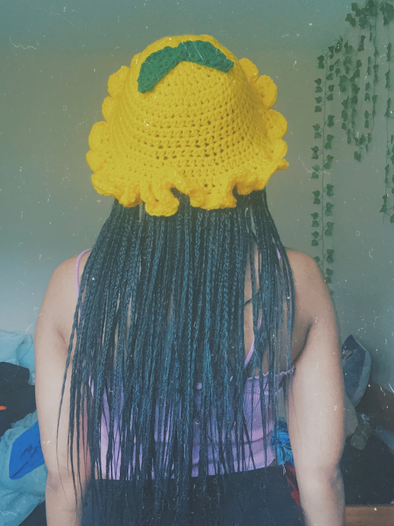 Pu&ograve; includere: Un cappello a secchiello di uncinetto giallo con foglie verdi in cima. Il cappello ha un bordo arricciato ed &egrave; indossato da una persona con lunghe trecce nere.