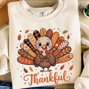 Könnte beinhalten: Cremefarbenes Sweatshirt mit einem Cartoon-Truthahn-Design. Der Truthahn hat einen bunten Schwanz und eine Fliege. Das Wort "Thankful" ist in einer Schreibschrift unter dem Truthahn geschrieben. Herbstblätter und Sterne umgeben den Truthahn.