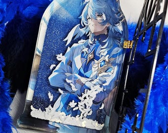 Op de afbeelding: Een blauw en wit anime-personage met lang wit haar is te zien in een heldere harsbedel. De bedel is omgeven door witte glitter en blauwe veren.