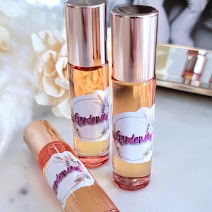 May include: Three glass perfume bottles with rose gold caps and labels that read "Gardenia" in pink script. The bottles are filled with a light orange liquid. The bottles are arranged on a white marble surface.