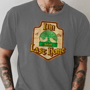 以下が含まれることがあります： 木製の看板に「Inn of the Last Home」と書いてあるグレーのTシャツ。看板には木と緑色の長方形の中に「Solace」という言葉が書かれている。