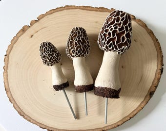 May include: Three brown and white morel mushroom shaped ceramic figurines with metal stems on a wooden surface.