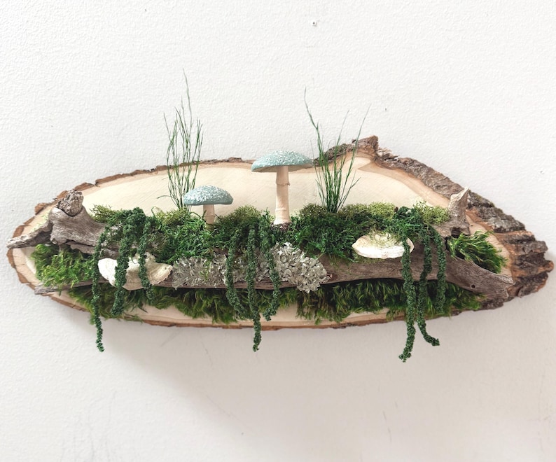 May include: A rustic wooden wall hanging featuring a natural wood slice with bark edges. It showcases a miniature forest scene with green moss, hanging vines, and two blue-green mushroom figurines. The piece is accented with sprigs of grass.