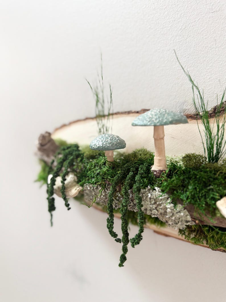 May include: A rustic wooden shelf featuring a miniature forest scene. Two light blue mushroom figurines with tan stems sit among green moss and hanging vines. Tall green grass blades add height to the natural decor.