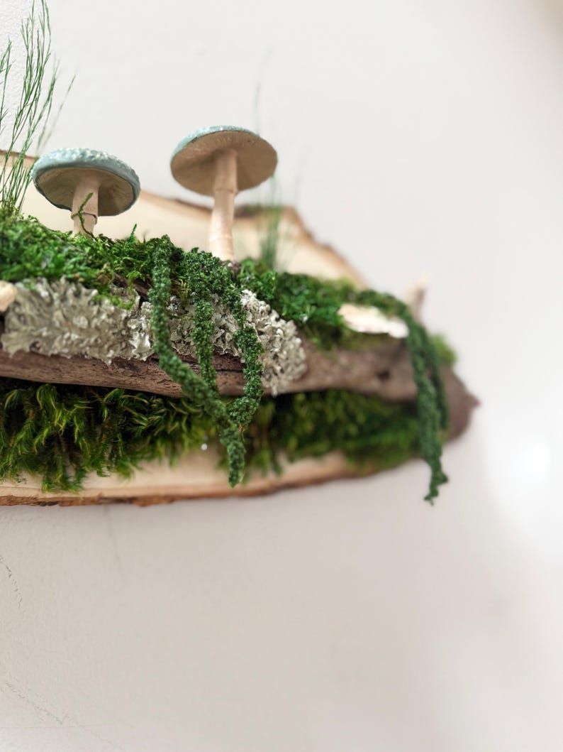 May include: A decorative wooden shelf featuring two mushroom sculptures with blue-green caps and tan stems. The shelf is adorned with green moss, lichen, and hanging greenery, creating a natural, woodland aesthetic. The shelf is made of a natural wood slice.