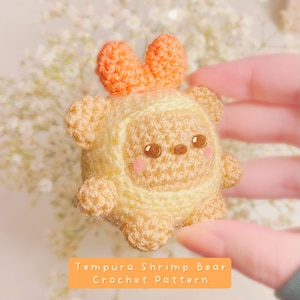 May include: A crocheted amigurumi toy bear in the shape of a tempura shrimp. The bear is yellow and brown with a heart-shaped orange bow on its head. The text "Tempura Shrimp Bear Crochet Pattern" is below the bear.