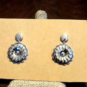 May include: A pair of silver-toned drop earrings featuring a smooth circular stud at the top connected to a larger, round, flower-shaped pendant with radiating petals and a central dot, all displaying a polished, reflective metallic finish.