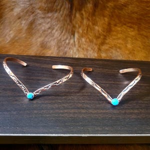 May include: Two V-shaped copper cuff bracelets, each featuring a small, round, bright turquoise stone set at the center point. The metal bands have a textured surface with geometric patterns etched along the outer edges of the open-ended design.