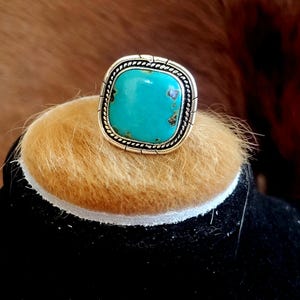 May include: A square turquoise ring with a silver and gold-tone setting. The turquoise stone is a vibrant blue-green color, and the ring has a detailed, braided border. The ring is displayed on a tan surface against a dark background.