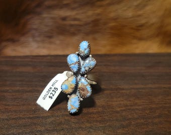May include: A silver ring featuring a cluster of teardrop-shaped turquoise stones with brown and blue hues. The ring has a unique, organic design. A tag with the text "GOLDEN HILL" is attached.