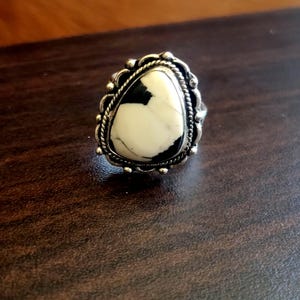 Può includere: Un anello in argento con una pietra bianca a forma di goccia caratterizzata da venature nere. La pietra è incastonata in una montatura decorativa in argento che presenta un bordo a corda intrecciata e piccole sfere d'argento lungo il perimetro.