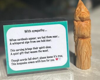 May include: A wooden carving of a bird, likely a cardinal, stands next to a card with the text "With sympathy..." and a message about remembrance. The carving is detailed with visible wood grain and a natural finish.