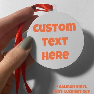 May include: A round white ornament featuring the orange text CUSTOM TEXT HERE, suspended by a red ribbon. Below the ornament, the text reads * SALMON VINYL FONT: LUCKIEST GUY. The ornament has a small loop at the top for hanging purposes.