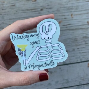 May include: A light blue sticker featuring a skeleton holding a margarita. The text on the sticker reads "Wasting away again in Margaritaville". The skeleton is white with black outlines.