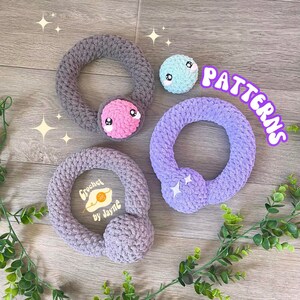 May include: Three crocheted rings with different colored yarn and embellishments. The rings are gray, purple, and light blue. The gray ring has a pink face with eyes and a mouth. The purple ring has two white stars. The light blue ring has a white face with eyes and a mouth. The text "PATTERNS" is written in white letters on a pink background. The text "Crochet by Jayne" is written in white letters on a yellow background.
