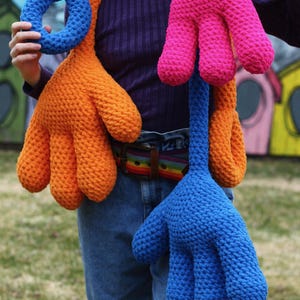 May include: A person wearing a blue shirt and jeans is wearing a long scarf made of three crocheted hand shapes. The hand shapes are orange, pink, and blue.