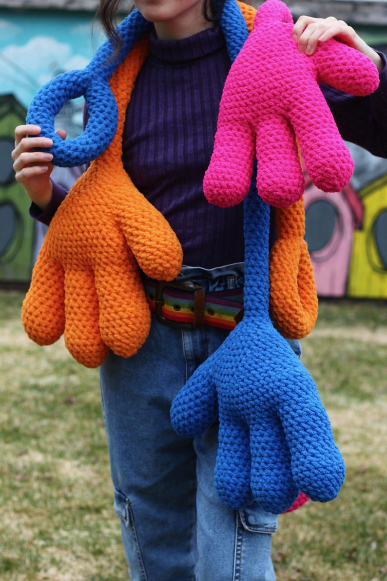 May include: A person wearing a blue shirt and jeans is wearing a long scarf made of three crocheted hand shapes. The hand shapes are orange, pink, and blue.