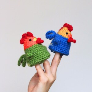 May include: Two handmade crochet finger puppets shaped like colorful roosters. One is green and red, the other is blue and yellow. The puppets are held up by a hand against a white background.